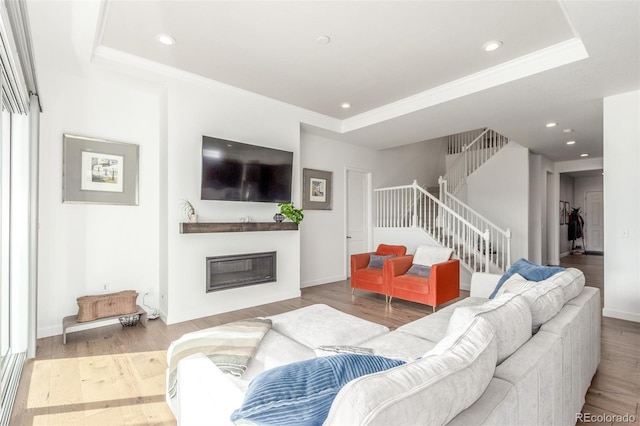 living room featuring a tray ceiling, a glass covered fireplace, wood finished floors, recessed lighting, and stairway