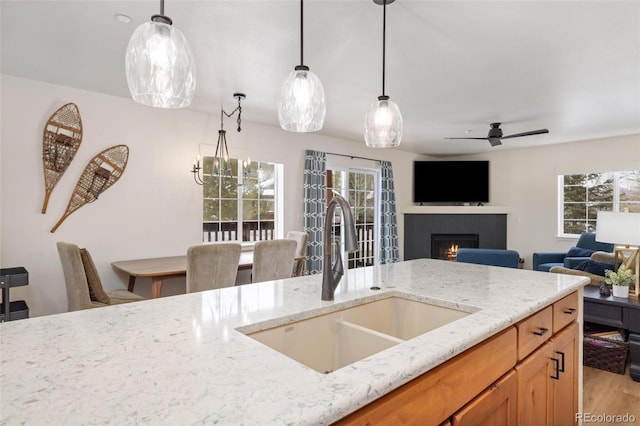 kitchen featuring a warm lit fireplace, light stone counters, light wood-type flooring, pendant lighting, and a sink