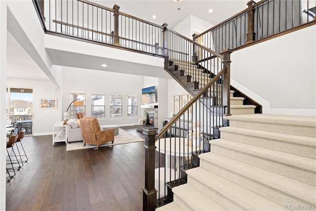 staircase featuring baseboards, a stone fireplace, recessed lighting, a towering ceiling, and wood-type flooring