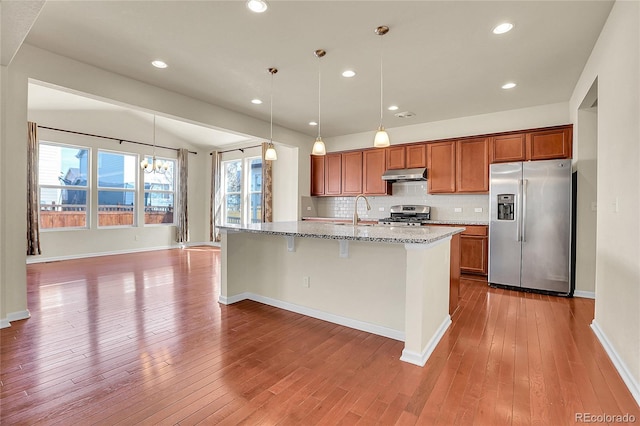 kitchen with brown cabinetry, wood-type flooring, light stone countertops, under cabinet range hood, and appliances with stainless steel finishes