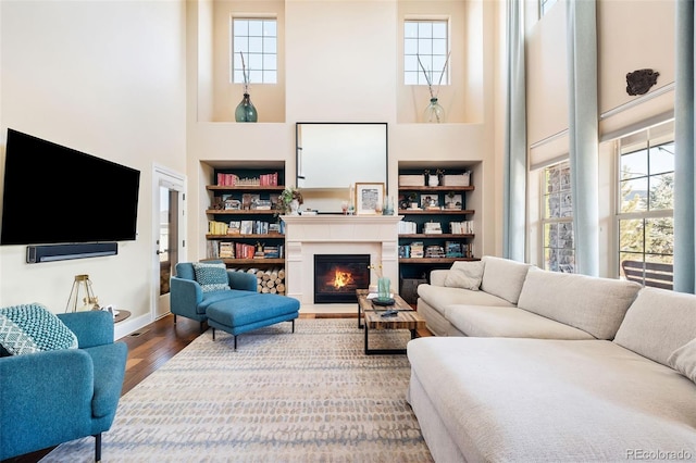 living area with a warm lit fireplace, baseboards, built in features, wood finished floors, and a high ceiling