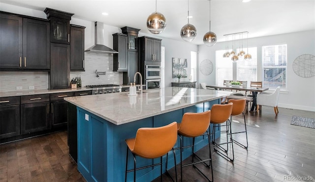 kitchen featuring dark wood-style floors, a kitchen bar, decorative backsplash, an island with sink, and wall chimney exhaust hood