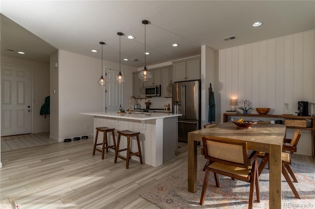 kitchen with light hardwood / wood-style floors, a center island with sink, gray cabinets, appliances with stainless steel finishes, and decorative light fixtures