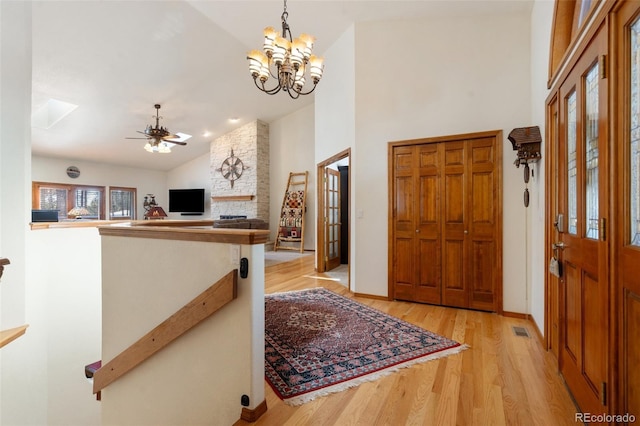 entryway with ceiling fan with notable chandelier, a skylight, high vaulted ceiling, a stone fireplace, and light wood-type flooring