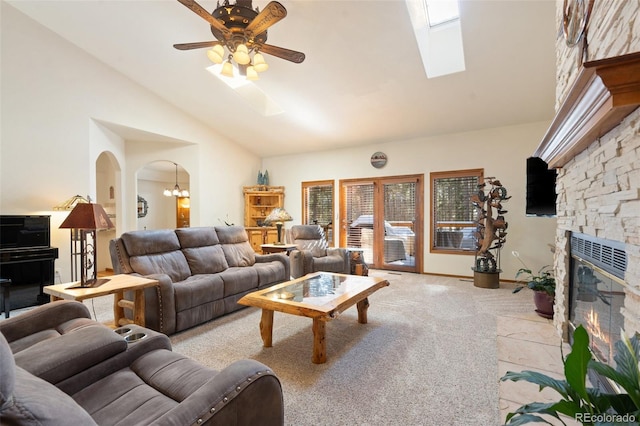 living room with light carpet, a stone fireplace, ceiling fan with notable chandelier, and vaulted ceiling with skylight