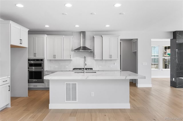 6683 Bridle Creek Pt, Castle Pines, CO 80108 - kitchen featuring wall chimney exhaust hood, stainless steel appliances, a kitchen island with sink, light stone counters, and light wood-style flooring