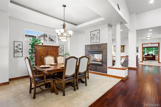 dining area featuring a tile fireplace, hardwood / wood-style floors, a notable chandelier, ornate columns, and a raised ceiling