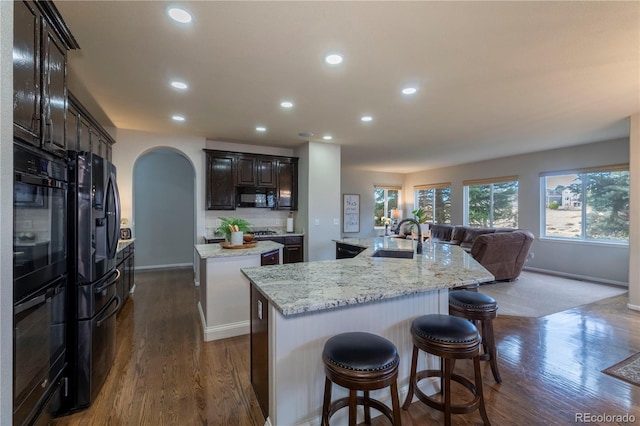 kitchen with sink, a kitchen island with sink, dark hardwood / wood-style floors, black appliances, and light stone countertops