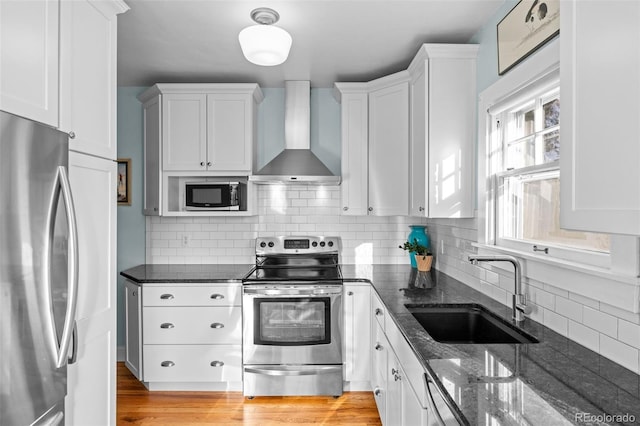 kitchen featuring a sink, white cabinets, wall chimney range hood, appliances with stainless steel finishes, and dark stone counters