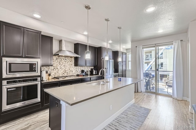 kitchen with appliances with stainless steel finishes, hanging light fixtures, sink, an island with sink, and wall chimney exhaust hood