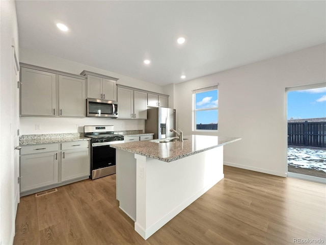 kitchen featuring gray cabinets, appliances with stainless steel finishes, an island with sink, light stone counters, and light hardwood / wood-style flooring