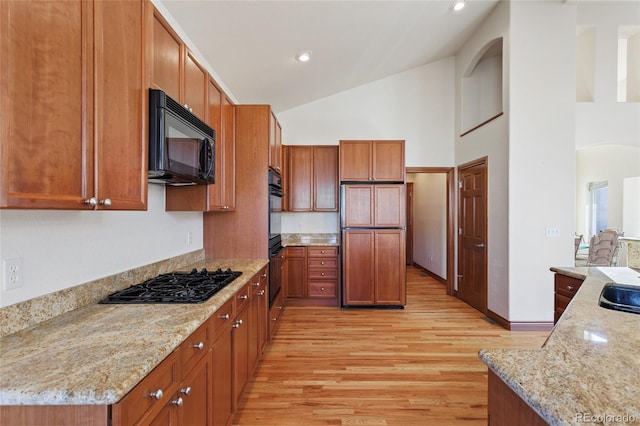 kitchen with high vaulted ceiling, black appliances, light hardwood / wood-style flooring, light stone counters, and sink