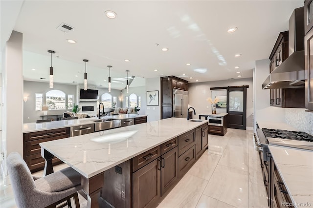 kitchen with a spacious island, wall chimney exhaust hood, sink, light stone counters, and hanging light fixtures