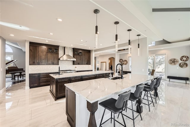 kitchen featuring wall chimney exhaust hood, sink, tasteful backsplash, decorative light fixtures, and a large island with sink