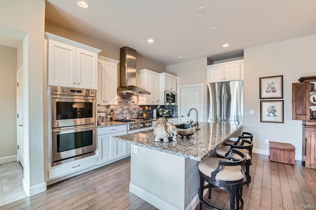 kitchen with decorative backsplash, light stone counters, stainless steel appliances, wall chimney range hood, and a sink