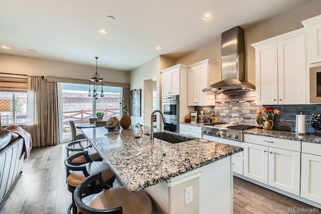 kitchen featuring stainless steel appliances, a sink, wall chimney range hood, decorative backsplash, and dark stone countertops