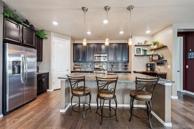 kitchen featuring stainless steel appliances, tasteful backsplash, a kitchen island with sink, light stone counters, and dark wood-style floors
