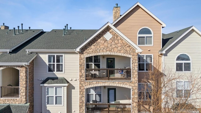 back of property with a shingled roof, a chimney, a balcony, and stone siding