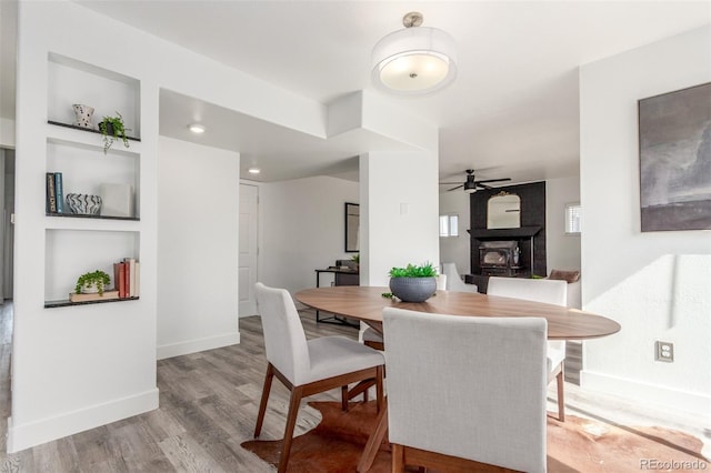 dining room with built in features, a fireplace, ceiling fan, wood finished floors, and baseboards