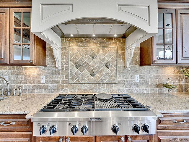 2131 Driver Ln, Erie, CO 80516 - kitchen featuring stainless steel gas stovetop, decorative backsplash, light stone counters, glass insert cabinets, and brown cabinetry
