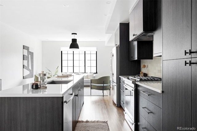 kitchen with wall chimney exhaust hood, decorative light fixtures, appliances with stainless steel finishes, a kitchen island with sink, and decorative backsplash