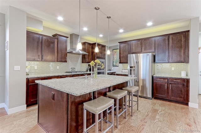 13713 Raritan Ln, Broomfield, CO 80023 - kitchen featuring appliances with stainless steel finishes, wall chimney range hood, light wood-type flooring, decorative backsplash, and a kitchen island with sink