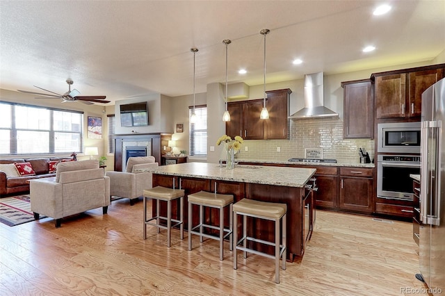 13713 Raritan Ln, Broomfield, CO 80023 - kitchen with wall chimney exhaust hood, stainless steel appliances, light wood-style floors, a breakfast bar, and a center island