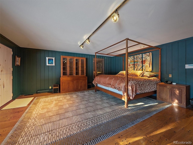 bedroom with wood-type flooring, baseboard heating, wooden walls, and rail lighting