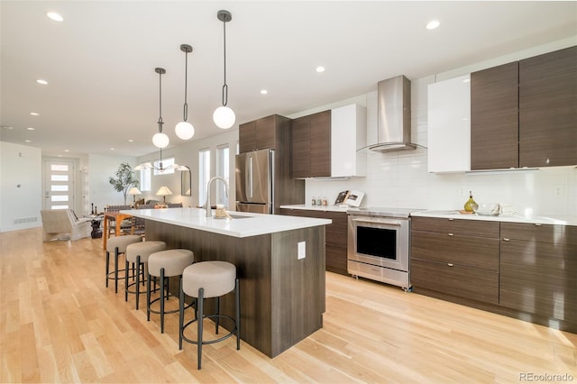 kitchen featuring sink, a center island with sink, appliances with stainless steel finishes, pendant lighting, and wall chimney range hood