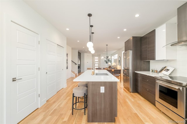 kitchen with sink, stainless steel appliances, dark brown cabinetry, an island with sink, and wall chimney exhaust hood