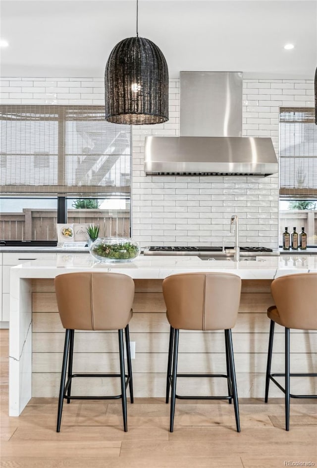 kitchen featuring light hardwood / wood-style flooring, hanging light fixtures, white cabinets, a kitchen bar, and wall chimney exhaust hood