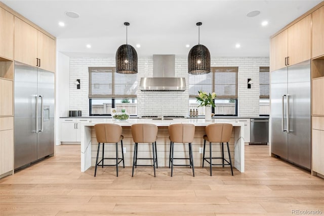 kitchen featuring wall chimney exhaust hood, decorative light fixtures, light brown cabinets, a kitchen island, and stainless steel appliances