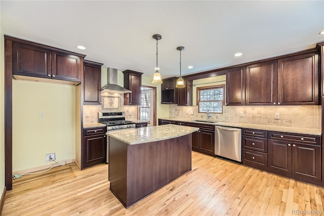 kitchen with wall chimney range hood, light hardwood / wood-style flooring, pendant lighting, a kitchen island, and appliances with stainless steel finishes