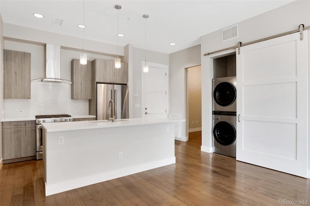 kitchen featuring wall chimney range hood, a barn door, stacked washer and dryer, a center island with sink, and appliances with stainless steel finishes