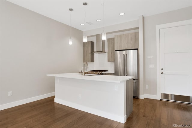 kitchen featuring high end fridge, sink, wall chimney range hood, hanging light fixtures, and an island with sink