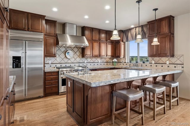 kitchen featuring wall chimney exhaust hood, light wood finished floors, tasteful backsplash, and high quality appliances