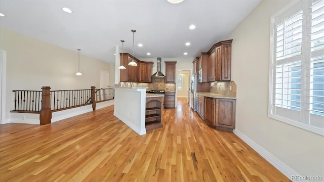 kitchen featuring wall chimney range hood, light hardwood / wood-style flooring, tasteful backsplash, light stone counters, and decorative light fixtures