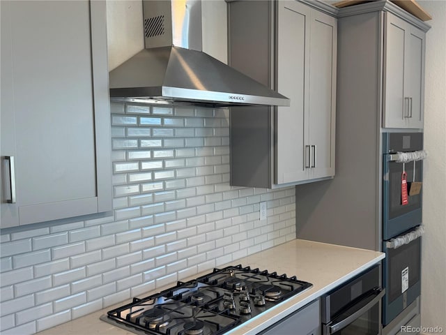 kitchen with double wall oven, gas stovetop, gray cabinetry, wall chimney range hood, and tasteful backsplash