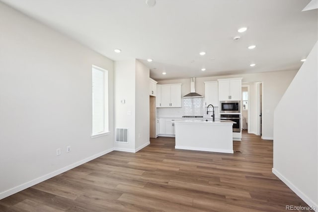 kitchen with built in microwave, white cabinetry, oven, a kitchen island with sink, and wall chimney range hood