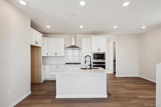 kitchen featuring white cabinets, oven, a center island with sink, and wall chimney range hood