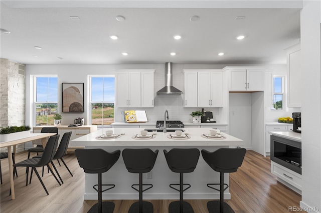 8863 Springsong Ln, Littleton, CO 80125 - kitchen featuring built in microwave, wall chimney exhaust hood, a center island with sink, white cabinetry, and light wood-style floors