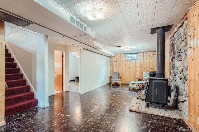 basement featuring wood walls, a drop ceiling, and a wood stove
