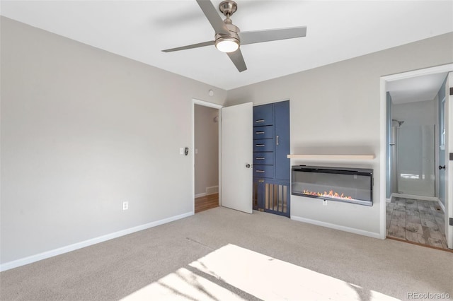 unfurnished bedroom with light colored carpet, a ceiling fan, baseboards, a closet, and a glass covered fireplace