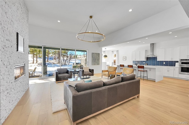 living area featuring recessed lighting, light wood-style flooring, a stone fireplace, a towering ceiling, and a chandelier
