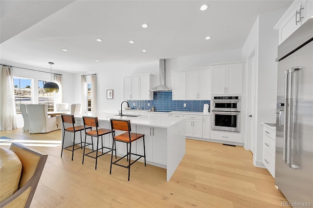 kitchen with appliances with stainless steel finishes, wall chimney range hood, a sink, a center island with sink, and tasteful backsplash
