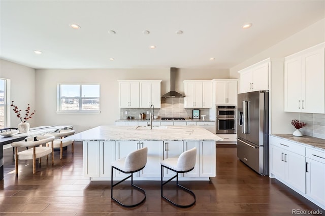 kitchen with light stone counters, wall chimney range hood, stainless steel appliances, and an island with sink