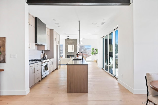 kitchen with dark countertops, wall chimney range hood, light wood-style flooring, stainless steel appliances, and a sink