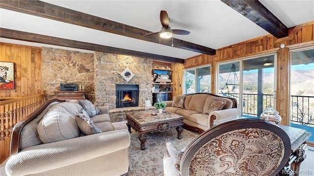 living room featuring ceiling fan, a stone fireplace, beam ceiling, and wood walls