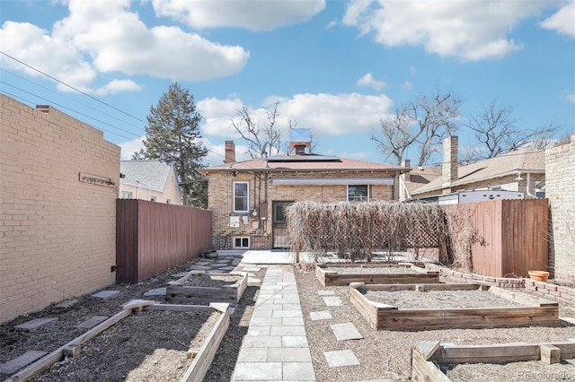 rear view of house featuring roof mounted solar panels, a vegetable garden, brick siding, fence private yard, and a chimney