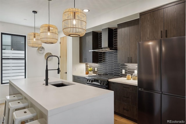 kitchen featuring stainless steel refrigerator, a kitchen island with sink, tasteful backsplash, gas stove, and wall chimney exhaust hood